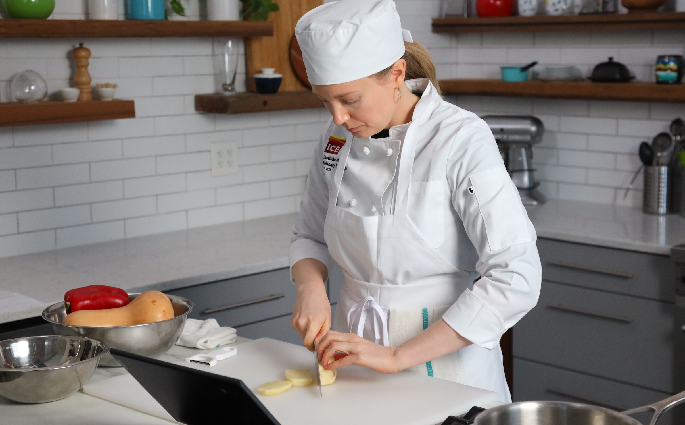 A student in chef whites with an ICE logo chops a vegetable for plant based culinary school online