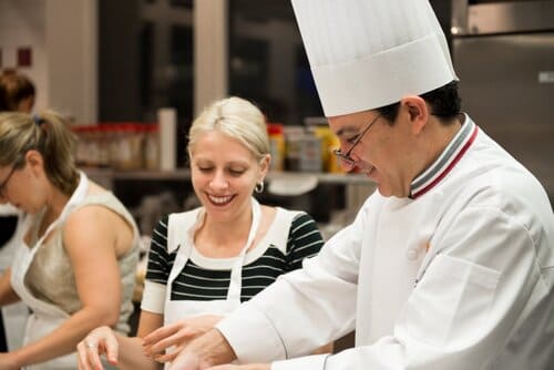 Woman smiles cooking beside chef in white coat and toque