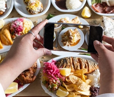 A person photographs a diverse spread of colorful dishes with a smartphone, showcasing vibrant salads, grilled fish, and garnished meals on a table.