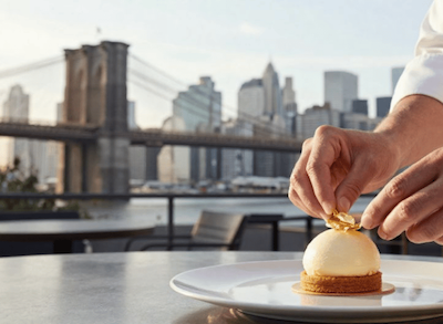 A chef is seen plating a dessert, highlighting attention to detail with Brooklyn Bridge in the background.