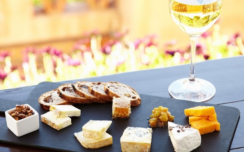 A slate cheese board arranged with various cheeses, sliced artisan bread, nuts, honey, and a glass of white wine on an outdoor table.