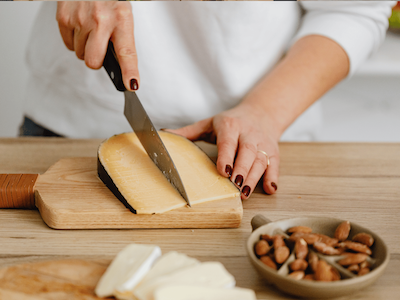 Close-up of a person with dark manicured nails slicing a wedge of Gouda cheese on a small wooden cutting board, with a small bowl of almonds in the foreground.
