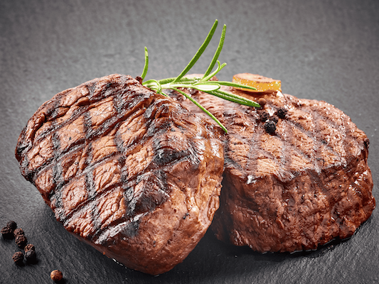Two grilled filet mignon steaks garnished with a sprig of rosemary and whole black peppercorns on a dark slate background.