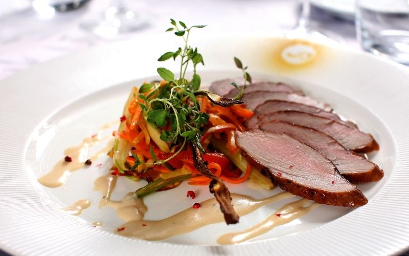 Slices of medium-rare roasted pork tenderloin arranged on a white plate alongside a colorful julienned vegetable salad topped with fresh herbs. A creamy pear sauce is drizzled around the dish, accented by small pink peppercorns.