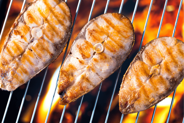 Three thick fish steaks cooking on a metal grill grate directly over bright, glowing orange flames.