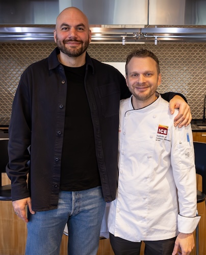 Chef Dan Giusti stands on the left. He is tall, bald, and has a dark beard. He wears a black open jacket over a black shirt and blue jeans. Chef Barry Tonkinson stands on the right. He wears a white chef's uniform jacket with an "ICE" logo.