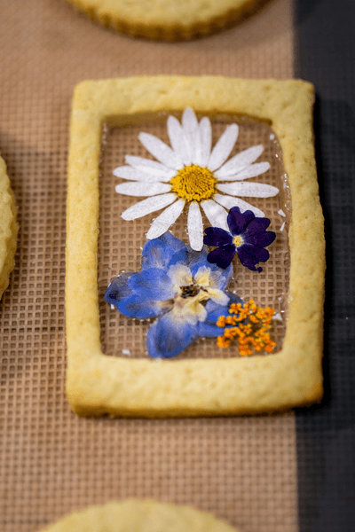 Rectangular cookie with pressed yellow flowers and green leaves in transparent jelly. Surrounding round cookies lie on a wooden surface.