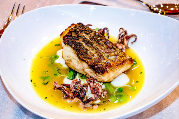A pan-seared white fish fillet with crispy, golden-brown skin served in a shallow white bowl with a yellow broth, green vegetables, and small pieces of grilled squid.