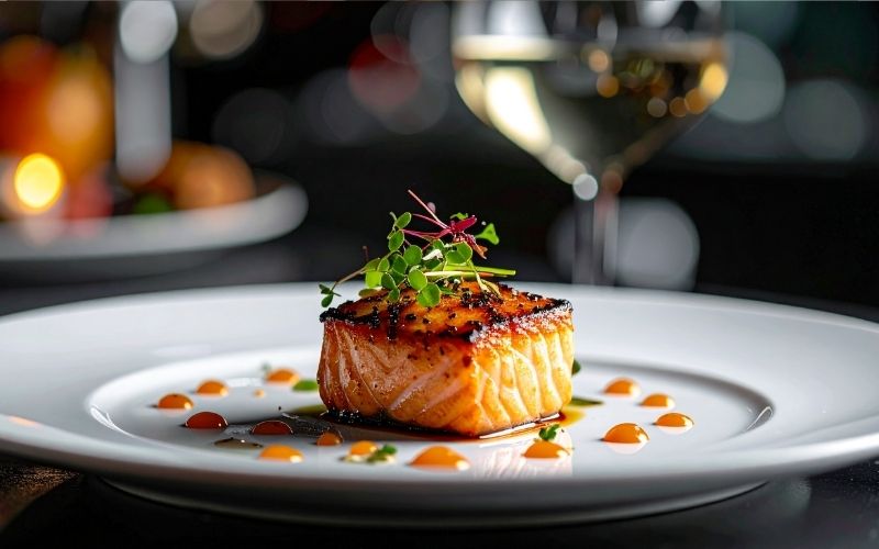 A close-up shot of a square piece of seared salmon filet with a caramelized glaze, served on a wide white plate and garnished with microgreens. 