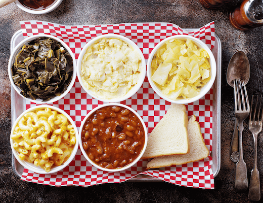 An overhead, top-down view of five classic barbecue side dishes in white bowls served on a metal tray lined with red and white checkered paper.