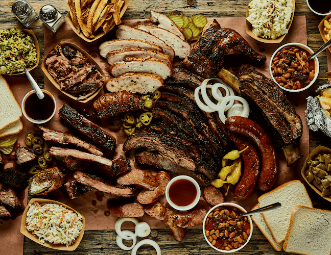 An overhead view of a large Texas barbecue spread on butcher paper, featuring sliced brisket, pork ribs, smoked turkey, sausages, pulled pork, baked beans, coleslaw and white bread.
