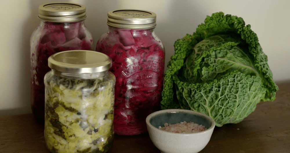A close-up photograph of homemade lacto-fermented cabbage on a wooden surface. Three glass jars filled with sauerkraut are visible: two tall Mason jars in the back contain vibrant pink or purple shredded cabbage, and one smaller jar in the foreground contains green shredded cabbage.
