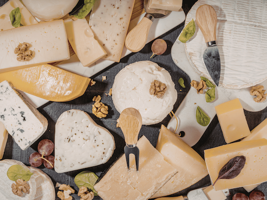 An overhead, flat lay view of an extensive assortment of artisanal hard, soft, and blue cheeses arranged on black and white serving boards, accented with walnuts, grapes, and small cheese knives.
