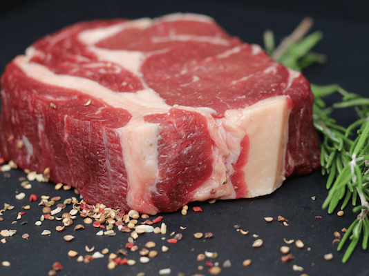 A thick, raw ribeye steak with white marbling on a black background, garnished with rosemary and peppercorns.