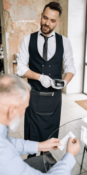A waiter wearing a vest, tie, and white gloves stands by a restaurant table holding out a portable card payment terminal to a seated customer reviewing a receipt.