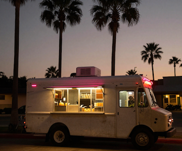 A white food truck parked at dusk with its service window illuminated, set against a background of tall palm trees silhouetted against a fading sunset sky.
