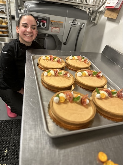 Chef Maried Arroyo smiling next to a tray of freshly decorated tarts on a stainless steel commercial kitchen counter.