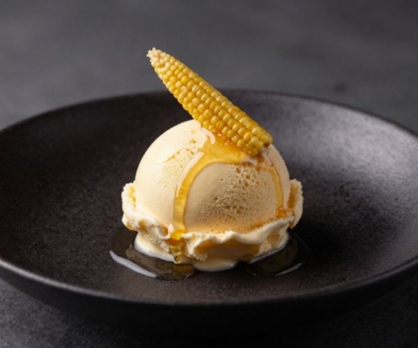 A scoop of pale yellow ice cream sits in a shallow, matte black bowl, drizzled with golden syrup. A small, whole baby corn is propped diagonally on top of the scoop.