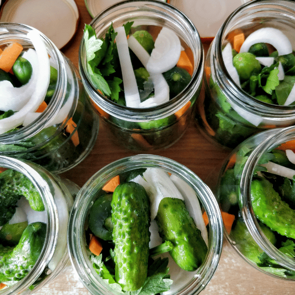Top-down view of six wide-mouth glass jars packed with fresh green pickling cucumbers, sliced white onions, orange carrot pieces, and leafy green herbs.