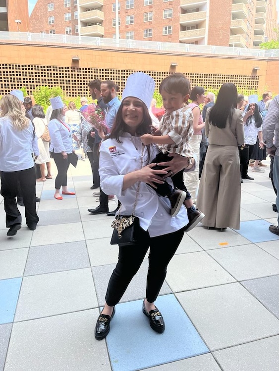 Maried Arroyo in her white chef uniform and hat holding her young son outdoors during a culinary school graduation event.