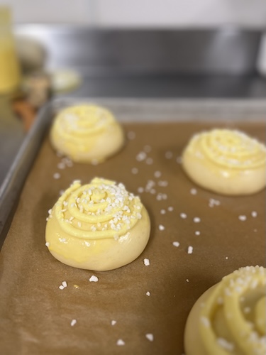 Unbaked pastry dough shaped into round sweet buns topped with a piped spiral custard ring and coarse pearl sugar on a lined baking sheet.