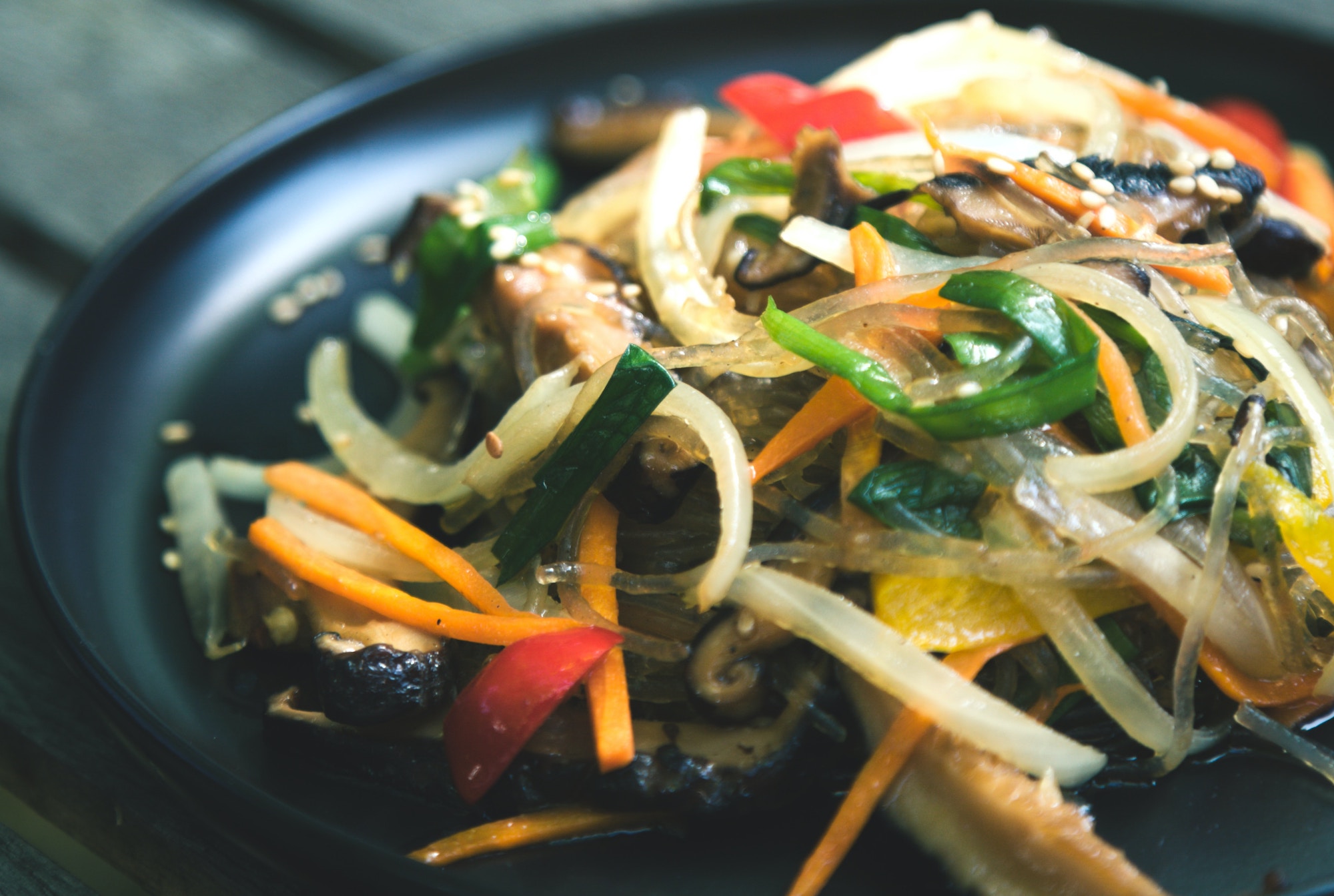 A colorful dish of glass noodles and vegetables sits on a dark colored plate