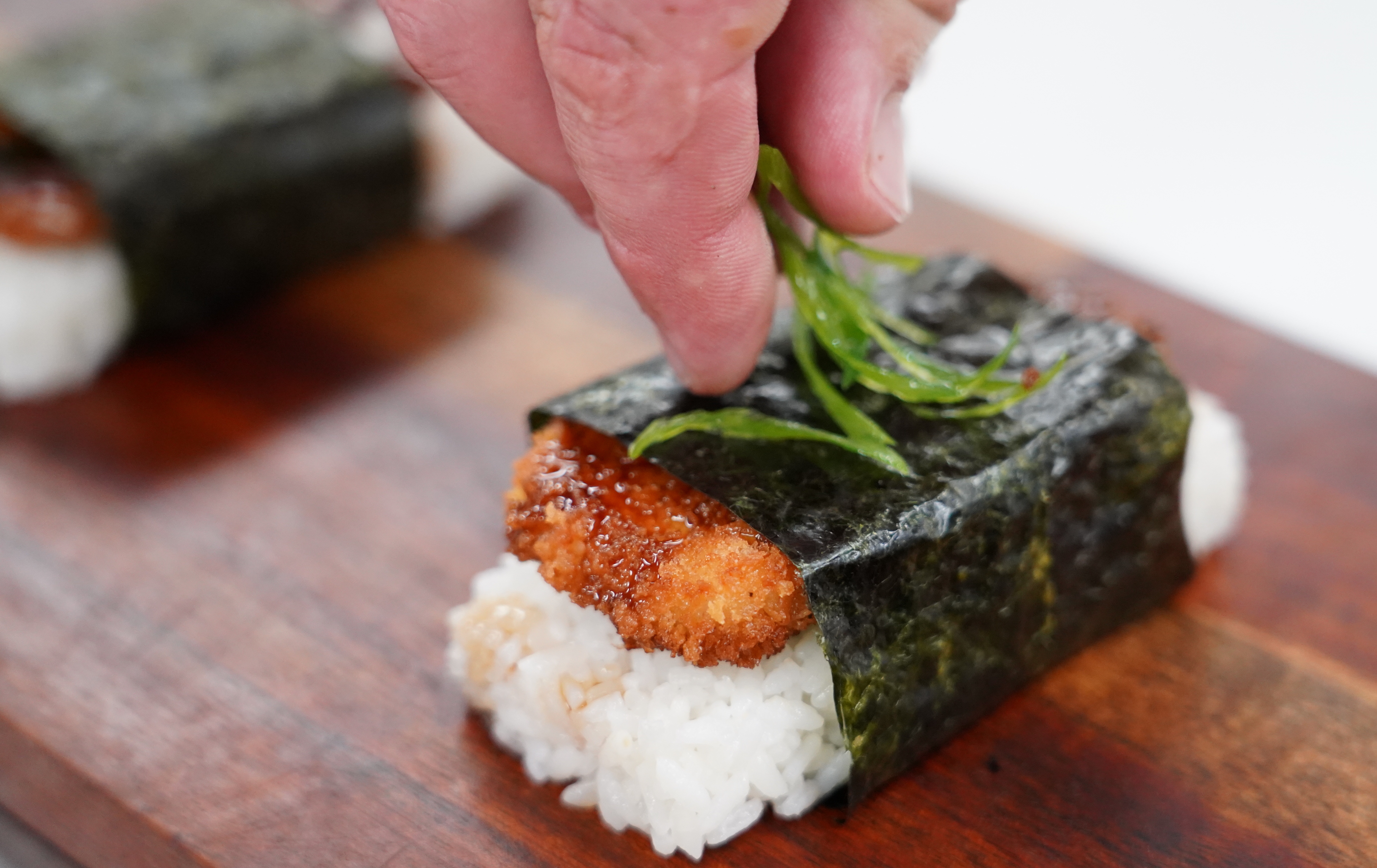 A piece of chicken katsu musubi on a brown wooden board is topped with sliced green onions