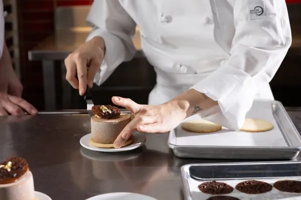 A pastry chef in a white uniform carefully places a delicate chocolate garnish onto a small, round mousse cake sitting on a metal kitchen counter.
