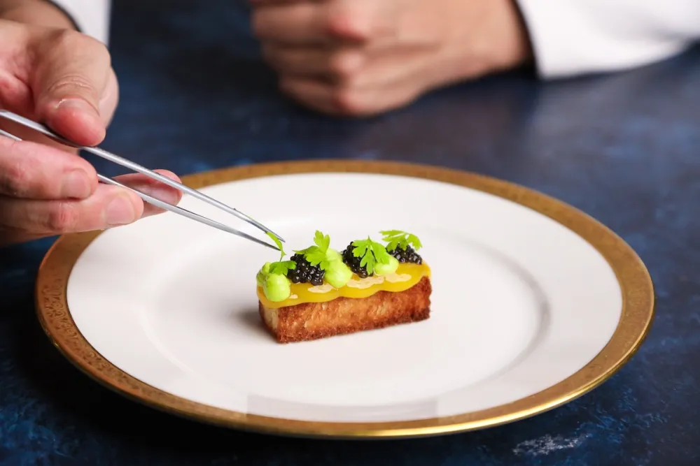 A close-up shot of a chef's hands using metal culinary tweezers to carefully place a small green herb garnish onto an elegant, rectangular hors d'oeuvre.