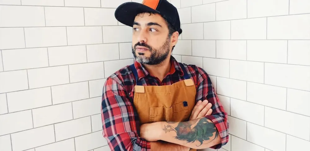 Chef David Viana stands in baseball cap, checkered shirt and leather apron in front of white tiled wall.