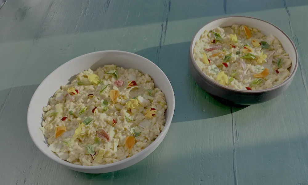 A creamy, light-colored risotto served in a white ceramic bowl, garnished with delicate, colorful edible flower petals and small green herbs. The bowl sits on a rustic, light-teal wooden surface illuminated by soft, natural sunlight from the upper left.