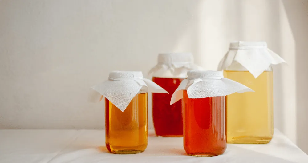 Four glass jars filled with liquids of varying amber and reddish shades are arranged on a white surface against a light background. Each jar is covered with a piece of white cloth secured around the rim.