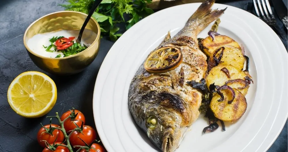 A whole grilled fish served on a white plate with charred lemon slices, grilled potatoes, and red onions, alongside a side sauce, a fresh lemon half, and cherry tomatoes.