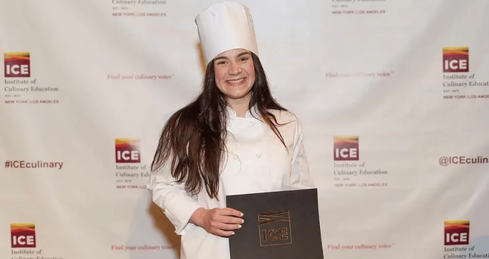 Maried Arroyo smiling in a white chef uniform and hat, holding her Institute of Culinary Education diploma in front of an ICE step-and-repeat backdrop.