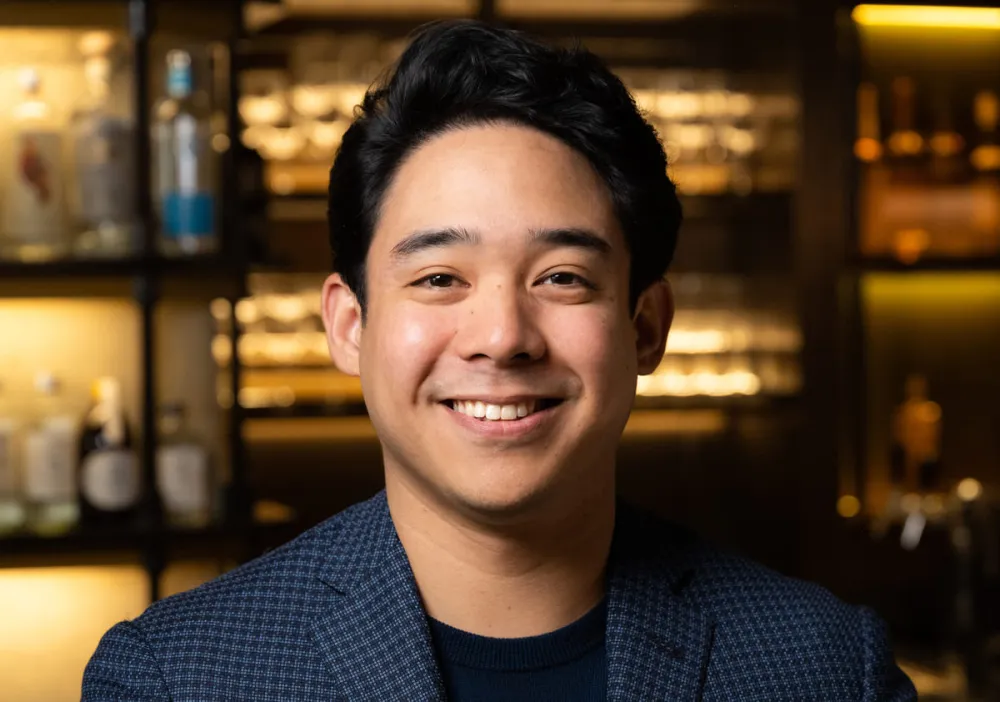 Jhonel Faelnar, Bev Director at NAE:UN Hospitality, smiles wearing navy blue blazer and black t-shirt standing in front of golden lit shelves full of beverages.