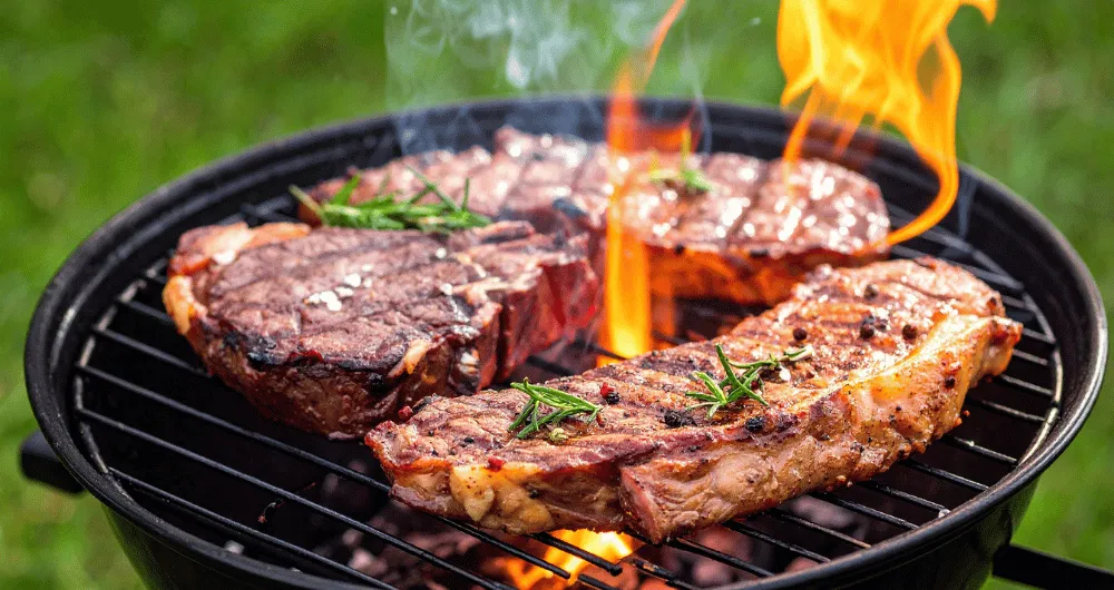Thick steaks grilling over an open flame on a charcoal barbecue, seasoned with peppercorns and fresh rosemary.
