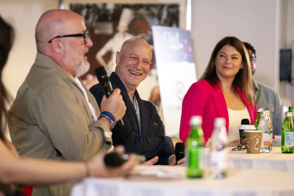 Food TV show host Andrew Zimmern speaks into microphone while Top Chef judges Tom Colicchio and Gail Simmons look on laughing.