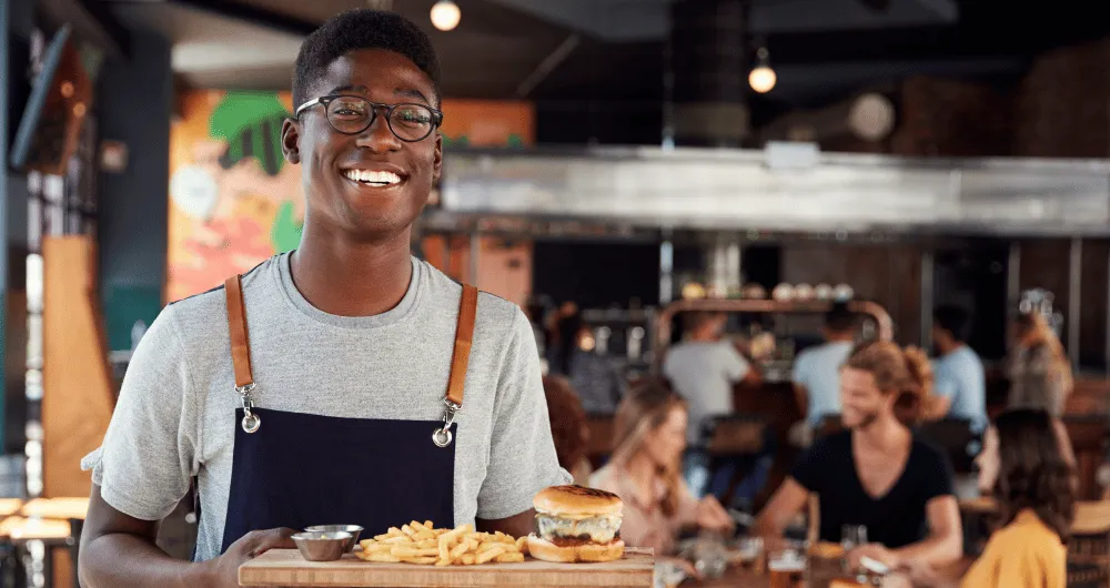 A server at a restaurant smiling with burgers and fries on a tray. 