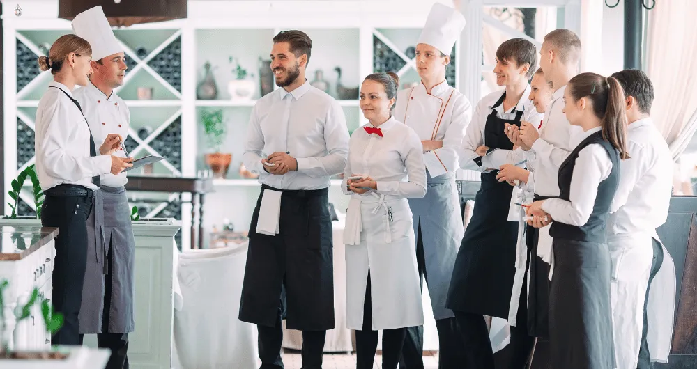 A group of restaurant staff, including chefs in white hats and servers in aprons, standing together and talking in a bright dining room.