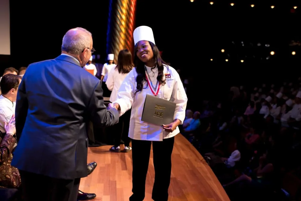 ICE culinary school graduate receives culinary arts diploma wearing chef toque and shaking hand of school president.