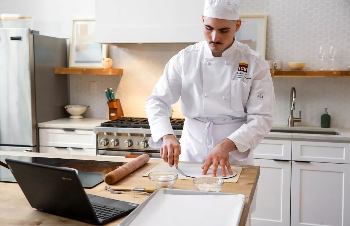 An ICE Online Pastry & Baking student in chef whites looks at a laptop while cutting a circle of dough