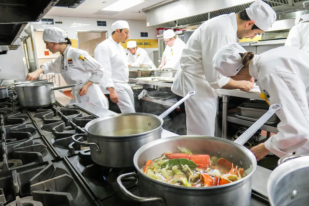 A bustling Institute of Culinary Education (ICE) commercial kitchen filled with students wearing white chef uniforms and matching caps.