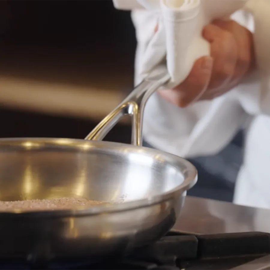 A chef in a white coat cooks on a stove with a stainless steel pan with sloped sides