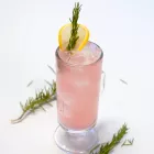 A pink drink garnished with rosemary and lemon on a white table