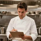 A chef in a restaurant writes notes on a clipboard, focused on organizing kitchen tasks and menu items.