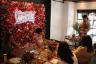 A group of people sit around a decorated wooden dinner table while ICE alumnus Jon Chandler prepares drinks behind a bar cart. In the background, a large wall covered in red and pink roses features a glowing neon Welch’s logo.