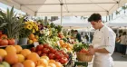 A young chef in a white uniform selects fresh vegetables and fruits at a vibrant outdoor market with colorful displays.