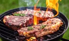 Thick steaks grilling over an open flame on a charcoal barbecue, seasoned with peppercorns and fresh rosemary.