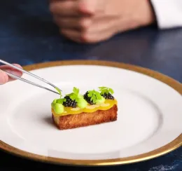 A close-up shot of a chef's hands using metal culinary tweezers to carefully place a small green herb garnish onto an elegant, rectangular hors d'oeuvre. 