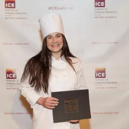 Maried Arroyo smiling in a white chef uniform and hat, holding her Institute of Culinary Education diploma in front of an ICE step-and-repeat backdrop.